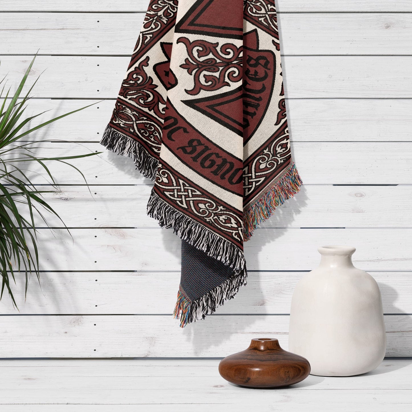 Woven throw blanket with Celtic knot pattern and fringe, draped on a white wooden wall.