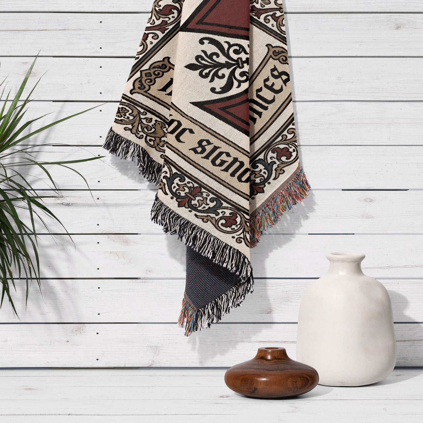 Woven blanket with fringe hanging on white wood wall next to vase.