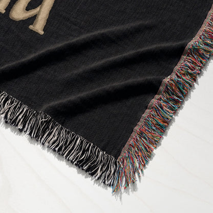 Black woven throw blanket with fringed edges and gold lettering.