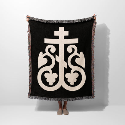 Black woven blanket with large cream-colored ornate cross symbol and fringed edges.