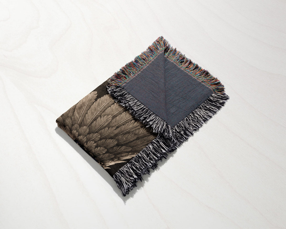 A folded woven blanket with a feather pattern and fringed edges rests on a white wooden surface.