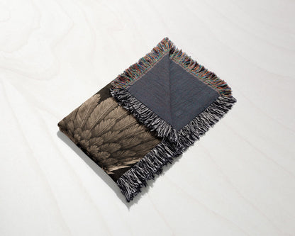 A folded woven blanket with a feather pattern and fringed edges rests on a white wooden surface.