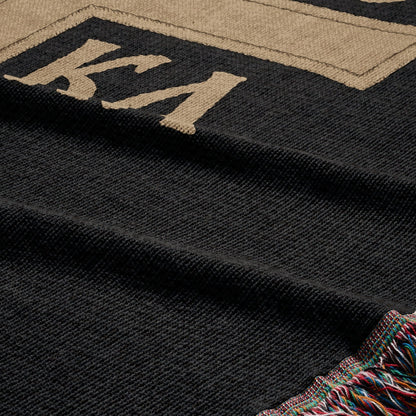 Black throw blanket with beige "KA" logo and colorful striped fringe.