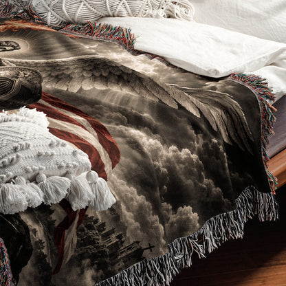 A woven throw blanket with a patriotic American flag and angel wing design lies on a bed with white pillows.