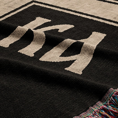 Black throw blanket with "KK" embroidered in beige and tasseled edges.