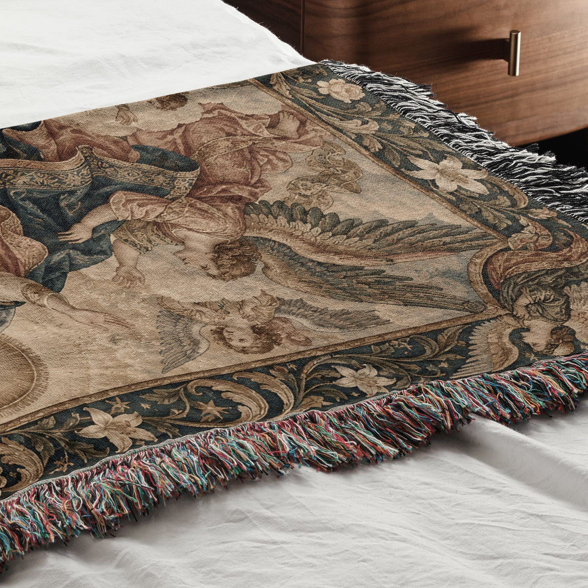 Woven throw blanket featuring a Renaissance art scene with winged figures, draped across a white bed.