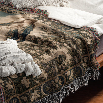 A woven tapestry-style throw blanket with a Renaissance art scene, fringed edges, draped over a bed.