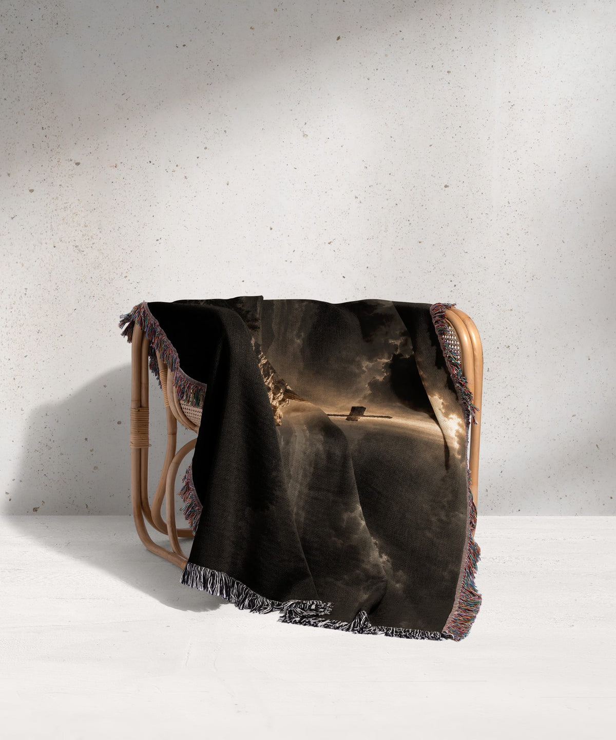 A woven blanket with a dramatic landscape design draped over a rattan armchair against a textured wall.
