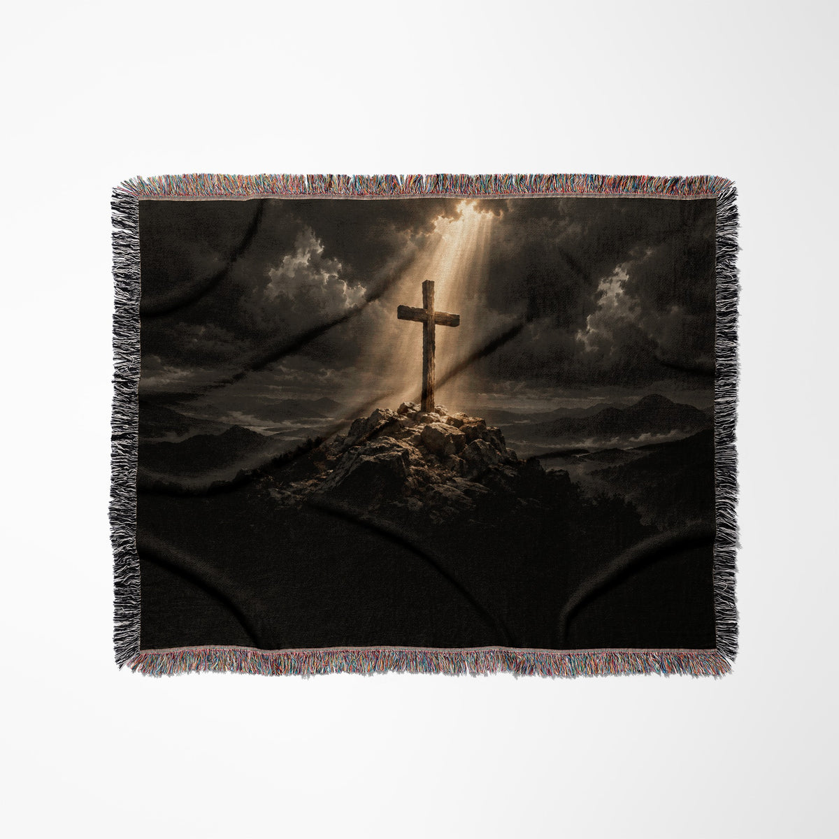 A woven throw blanket displays a wooden cross on a rocky hilltop beneath dramatic skies and sunlight beams.