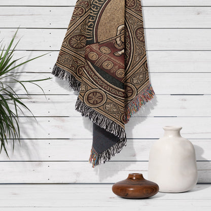 Woven blanket with intricate circular pattern and tassels, draped on white wall.