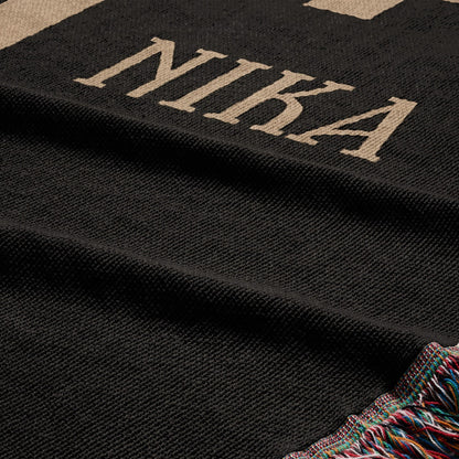 Black woven blanket with beige "NIKA" text and fringed edge.