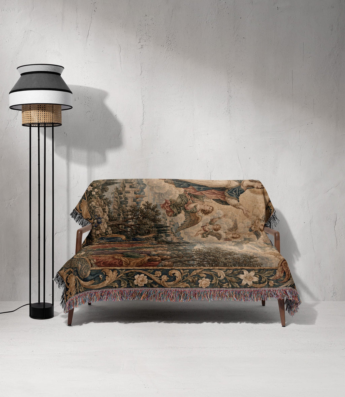 A vintage-style tapestry throw blanket with a Renaissance scene and fringed edges covers a wooden sofa next to a tall standing lamp against a textured white wall.