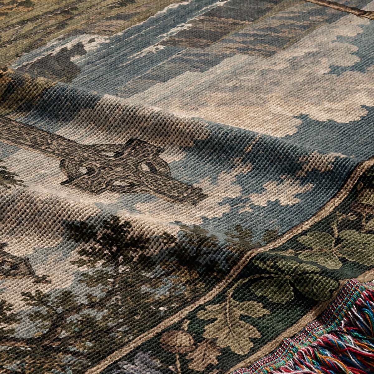 A folded woven throw blanket featuring a landscape design with a cross and fringe trim.