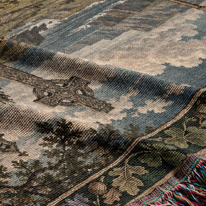 A folded woven throw blanket featuring a landscape design with a cross and fringe trim.
