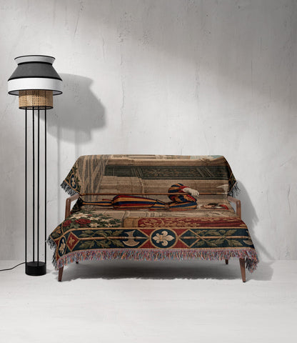 A modern wooden sofa draped with a large, Renaissance-style tapestry throw blanket featuring a standing guard, set against a concrete wall next to a tall, layered floor lamp.
