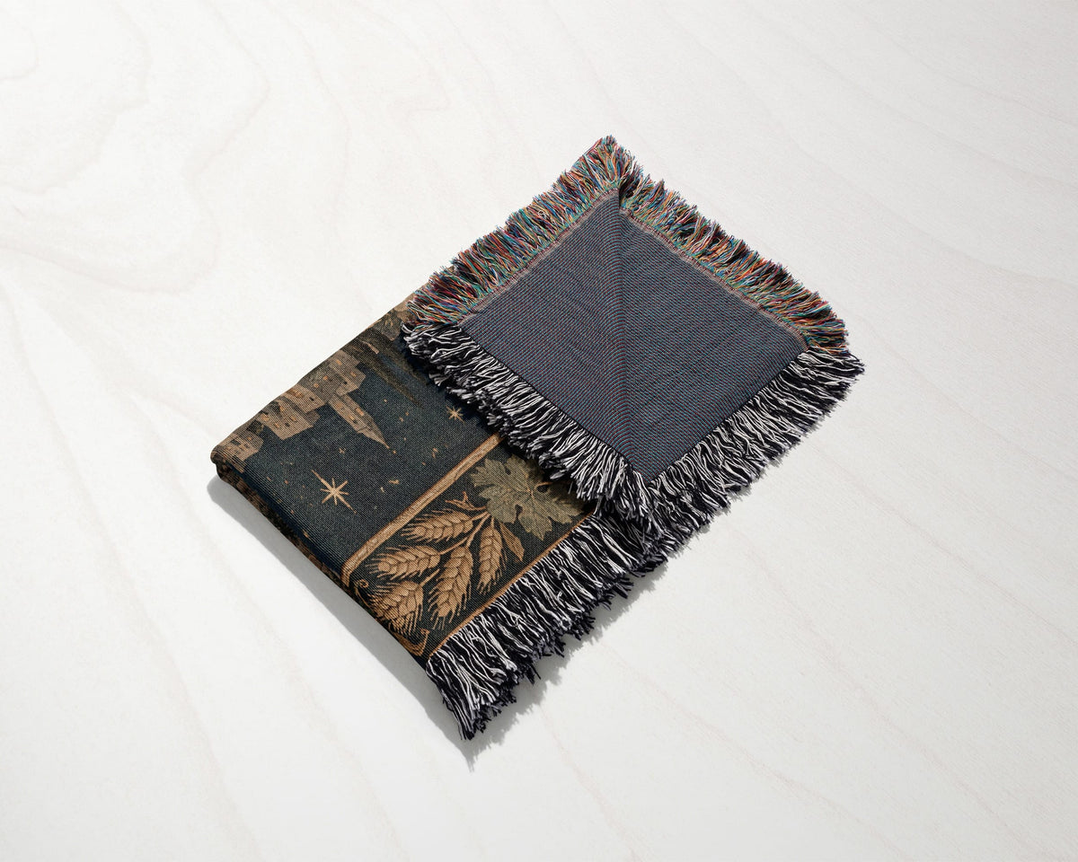 A folded woven blanket with a dark celestial and botanical pattern, featuring fringed edges on a light surface.