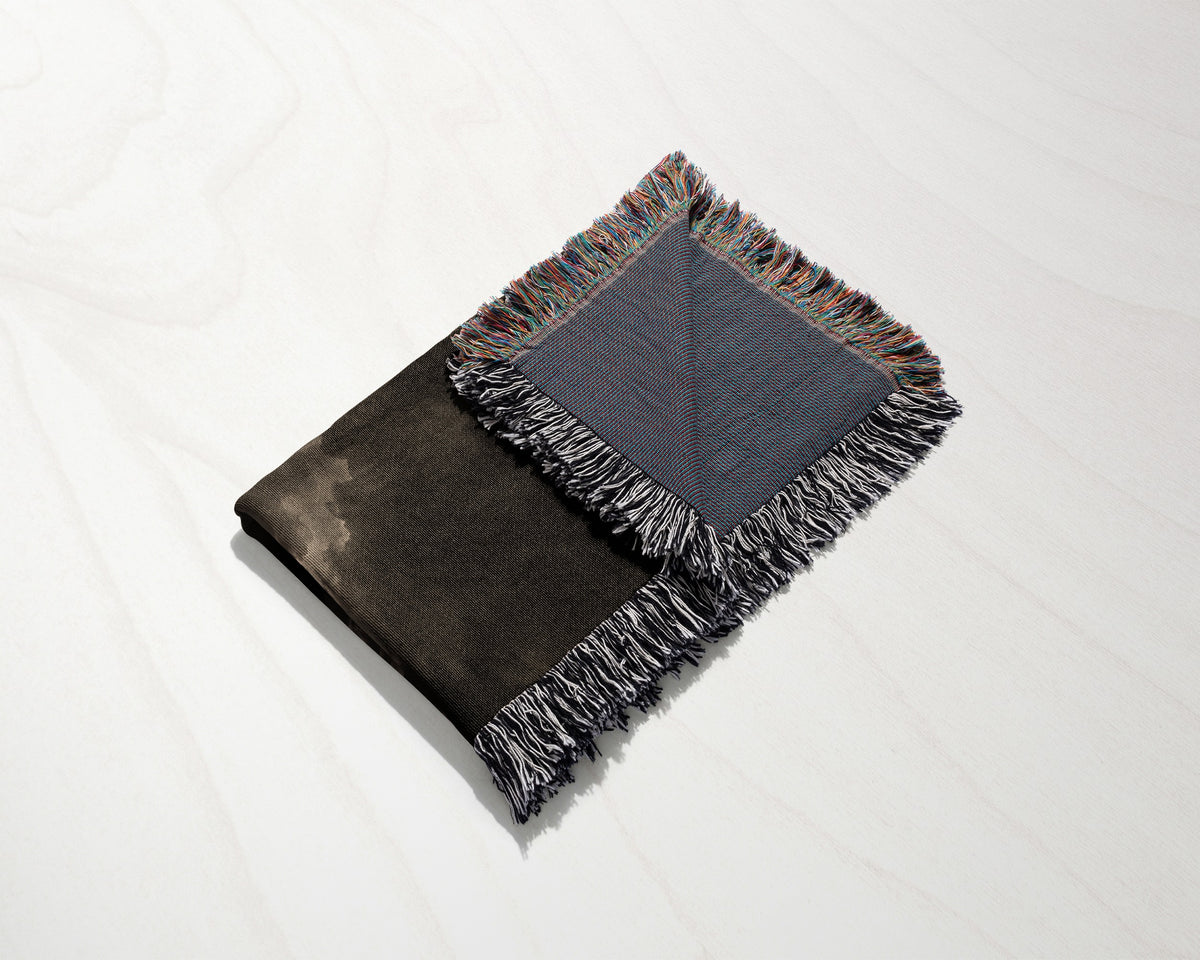A folded black and blue woven throw blanket with fringe rests diagonally on a white wooden surface.