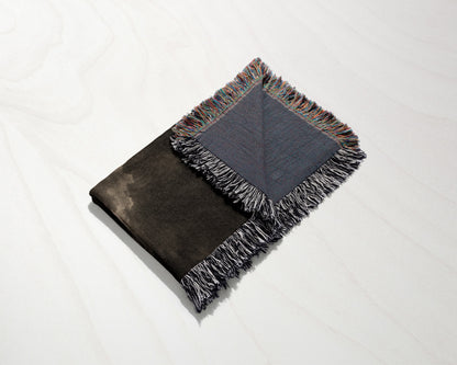 A folded black and blue woven throw blanket with fringe rests diagonally on a white wooden surface.