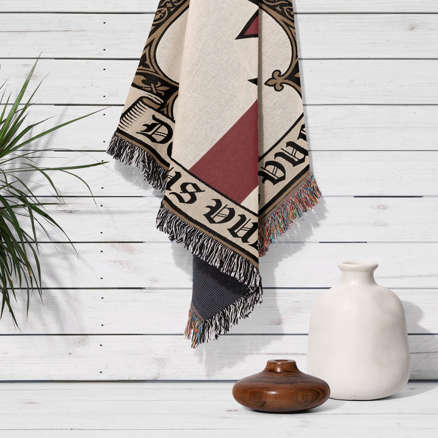 Cream and maroon throw blanket with fringe hanging on white wall, with vase and plant.