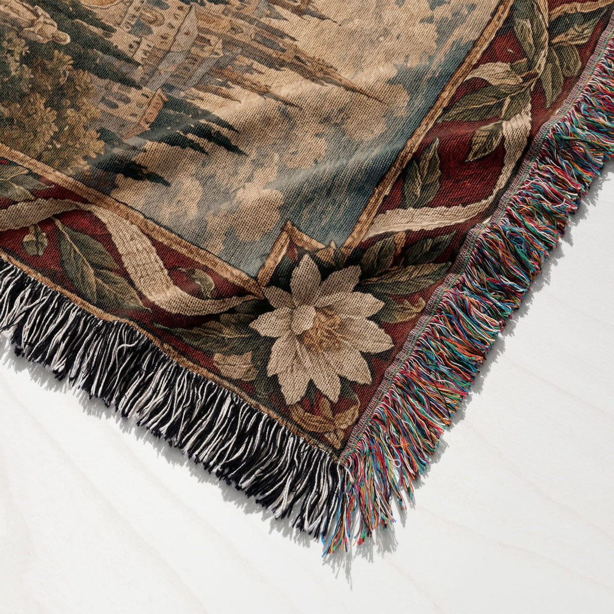 Folded woven tapestry throw blanket featuring a classic European landscape scene and decorative floral border with fringe.