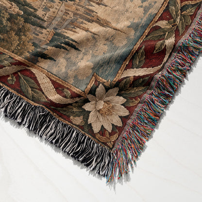 Folded woven tapestry throw blanket featuring a classic European landscape scene and decorative floral border with fringe.