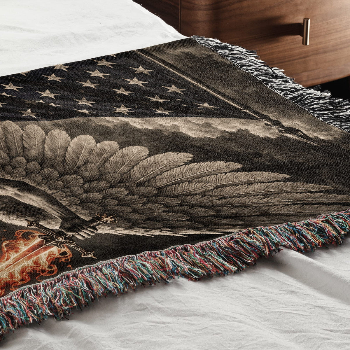 A woven blanket featuring an American flag, feathered wings, and a fiery sword resting on a bed.