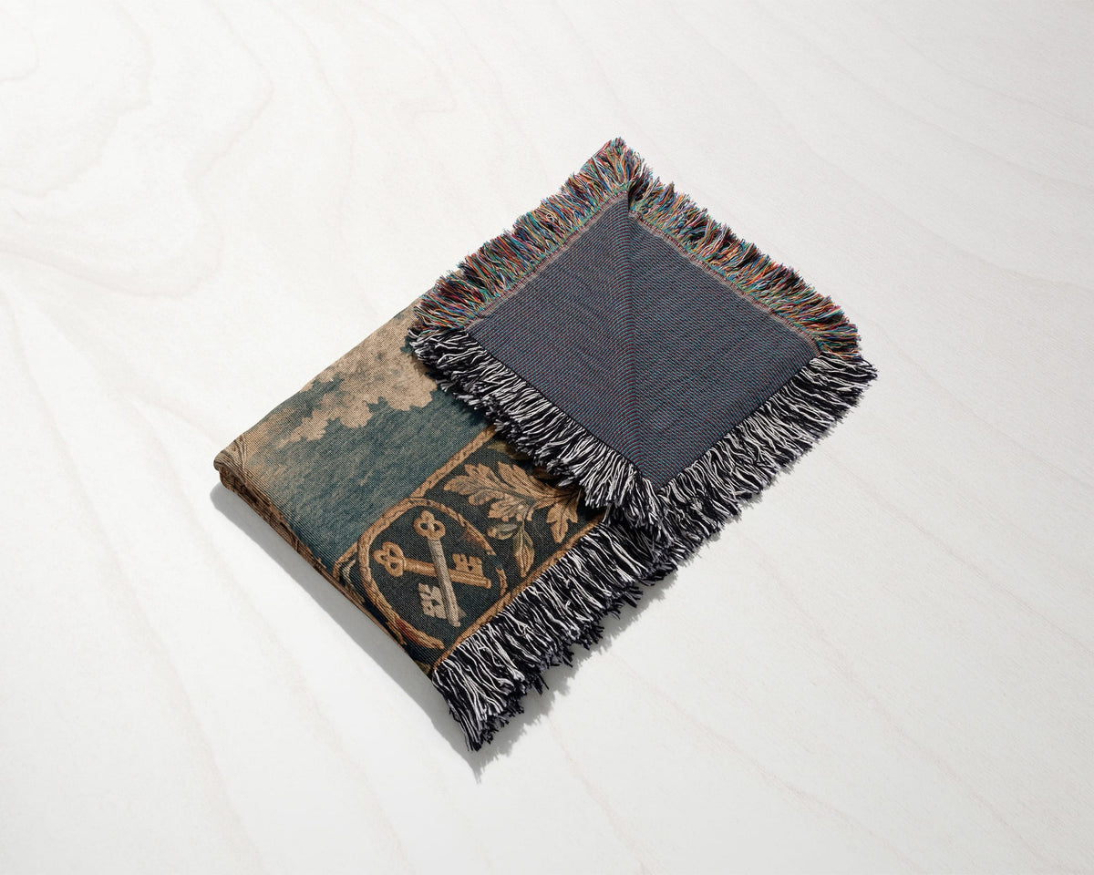 A folded woven tapestry throw blanket with a medieval design featuring crossed keys and fringe edges.