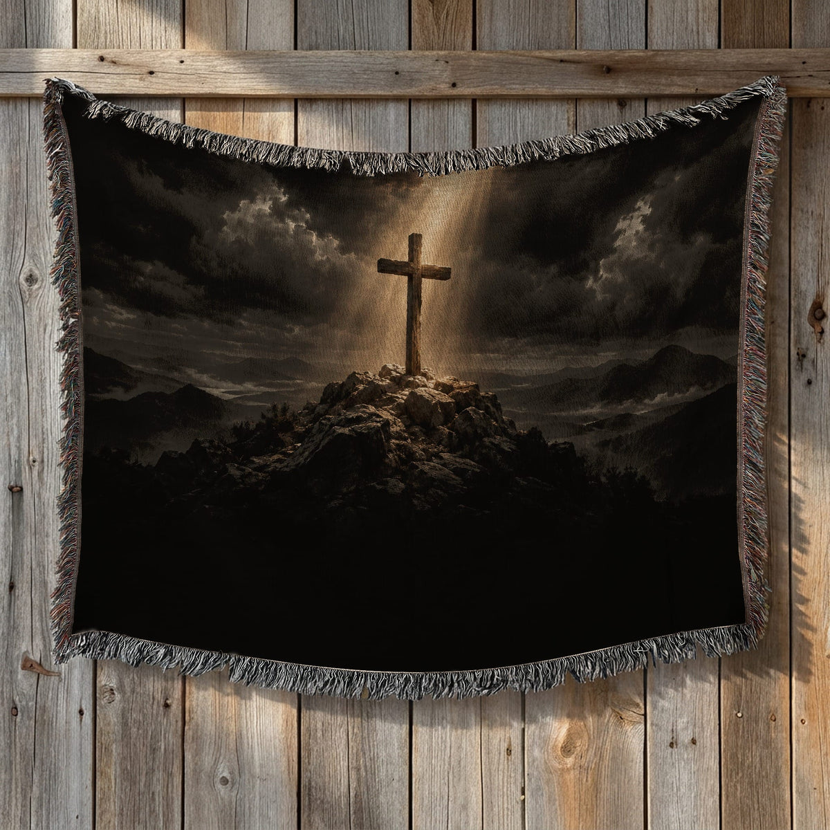 Woven blanket featuring a wooden cross atop a rocky hill under a cloudy, sunlit sky, hanging against a wooden wall.