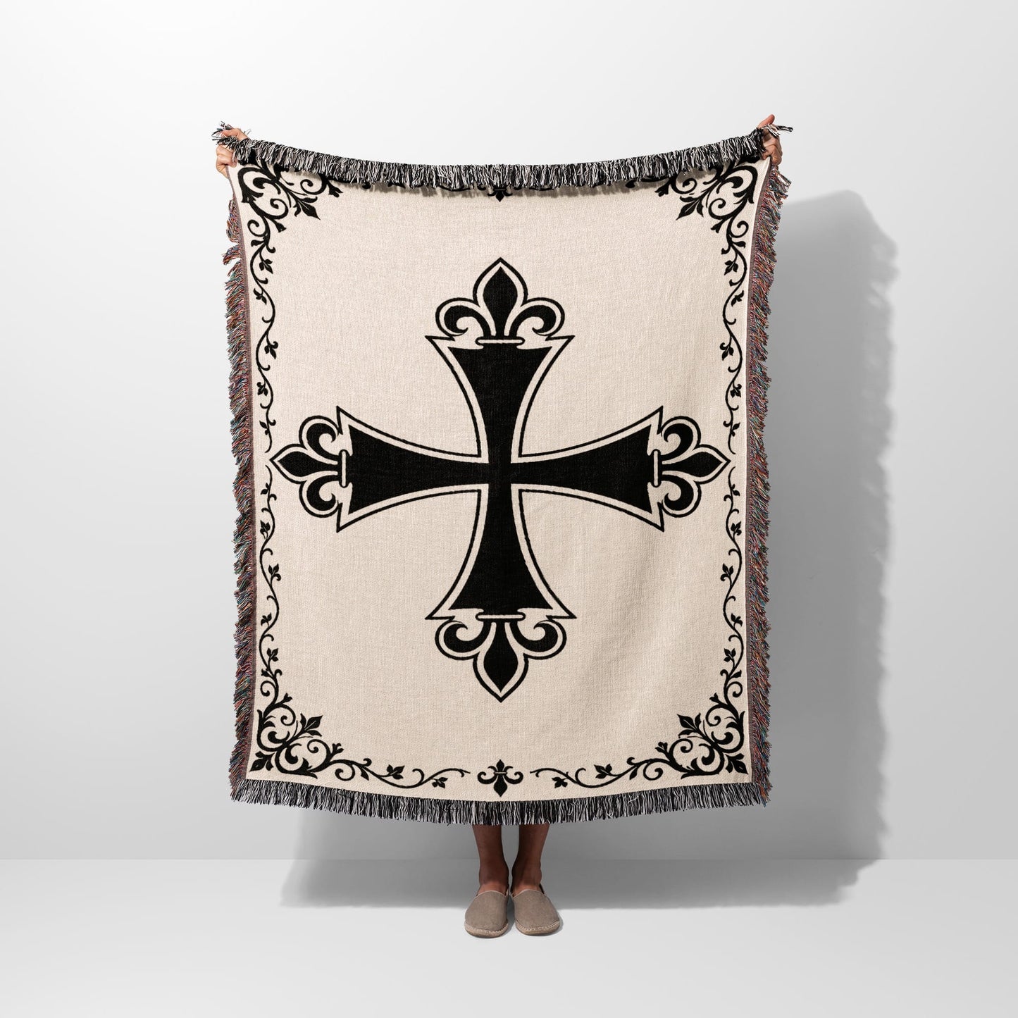 Beige throw blanket with black ornate cross and fringed edges.
