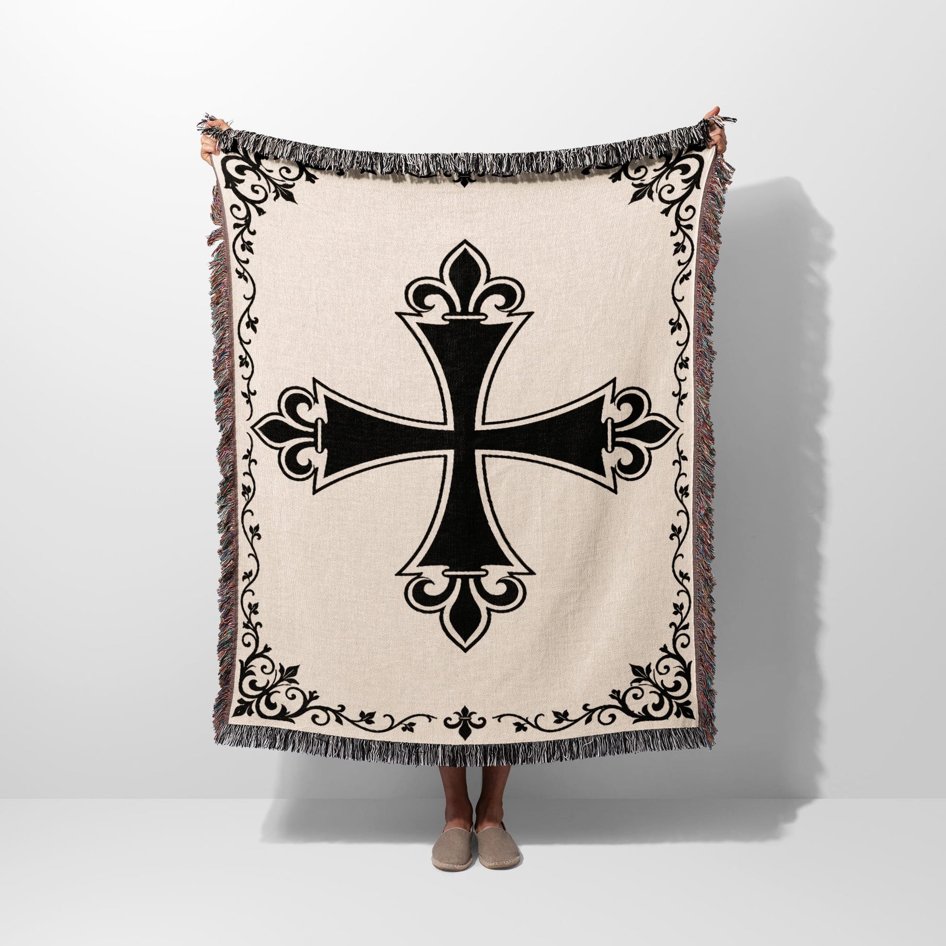 Beige throw blanket with black ornate cross and fringed edges.