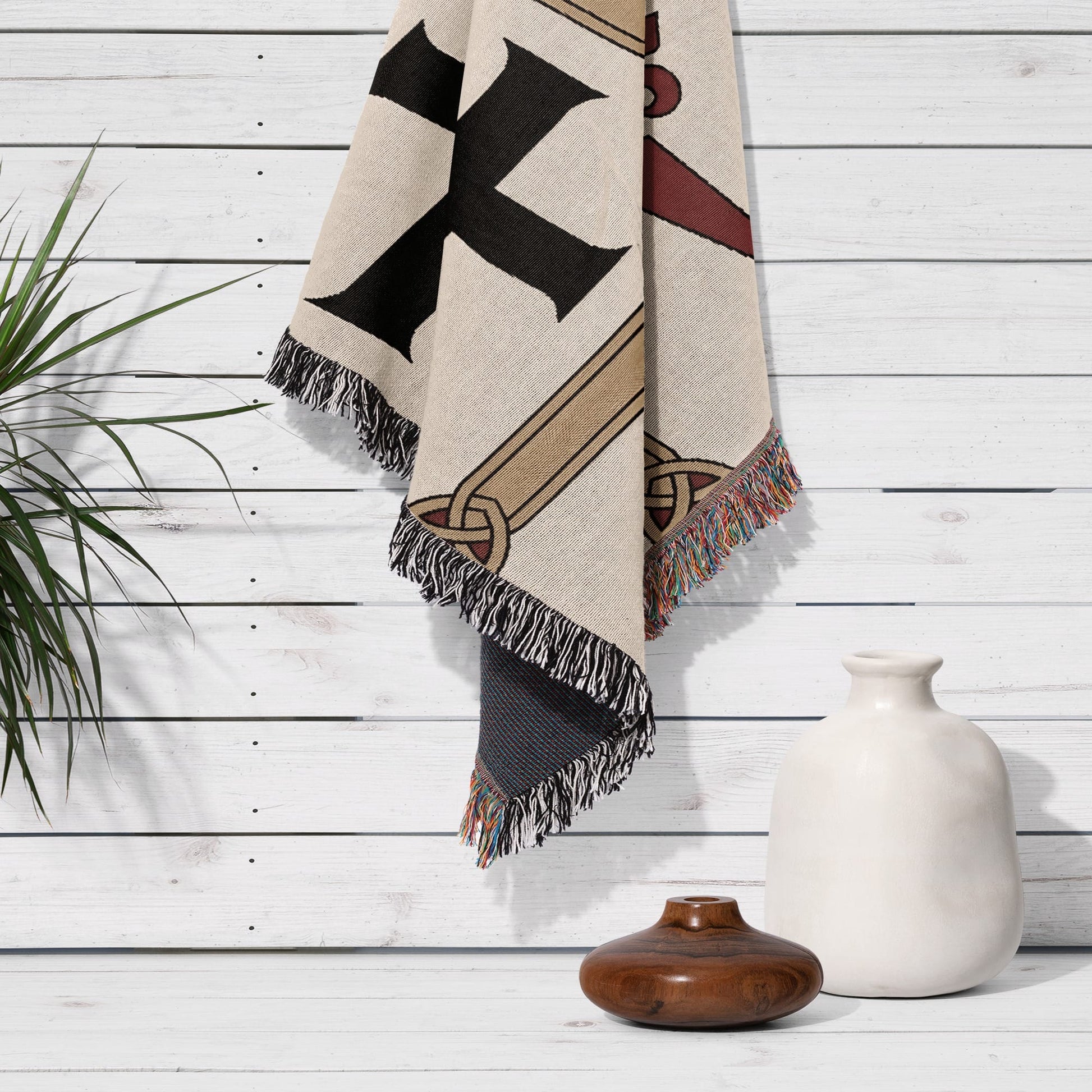 Beige throw blanket with a black and gold Celtic knot design, draped over a white wooden wall.