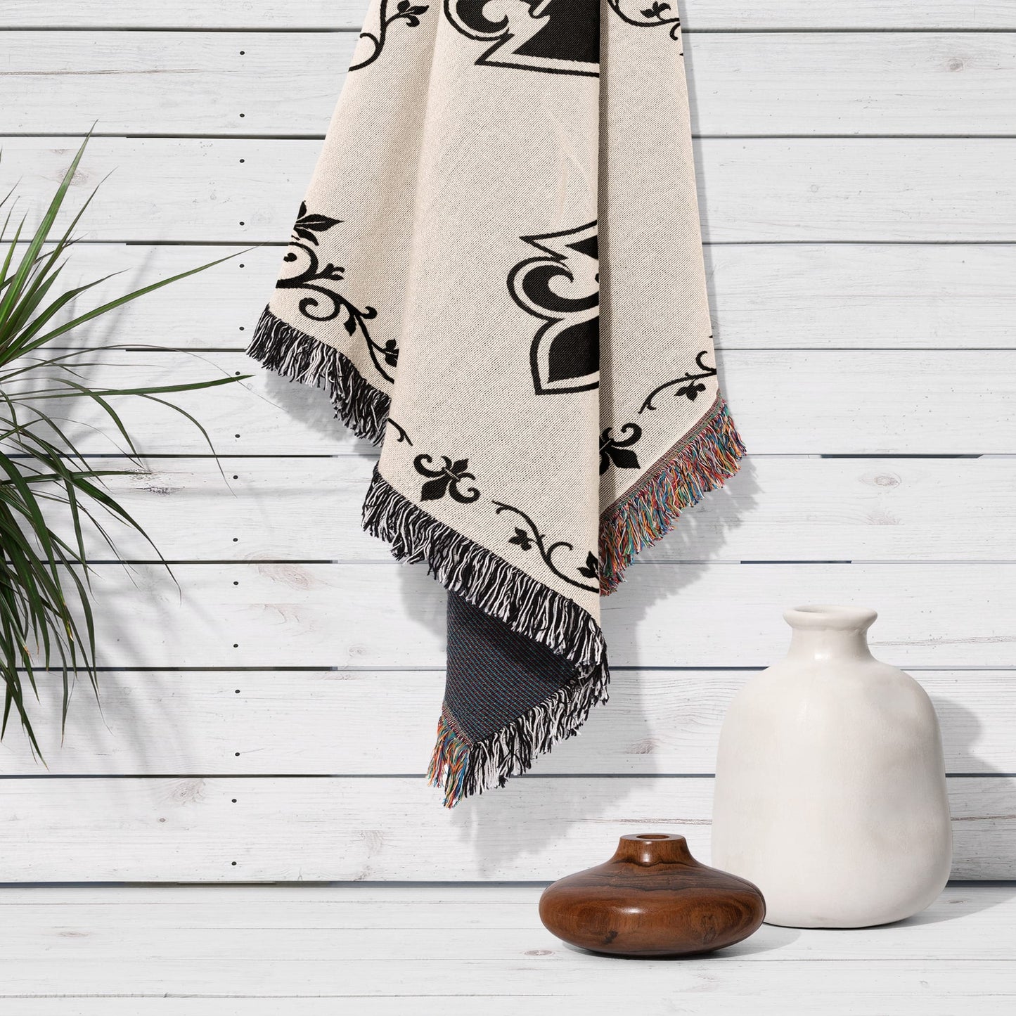 Cream blanket with black floral pattern and tasseled edges hangs on a white wooden wall.