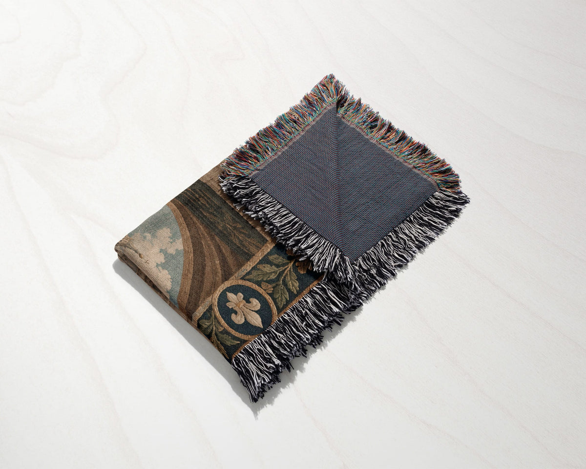 A folded tapestry throw blanket with fringe rests on a light wooden surface.