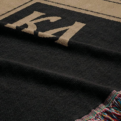 Black woven blanket with tan "KA" logo and fringed edge.