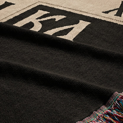 Black woven blanket with multicolor fringe and large white text.