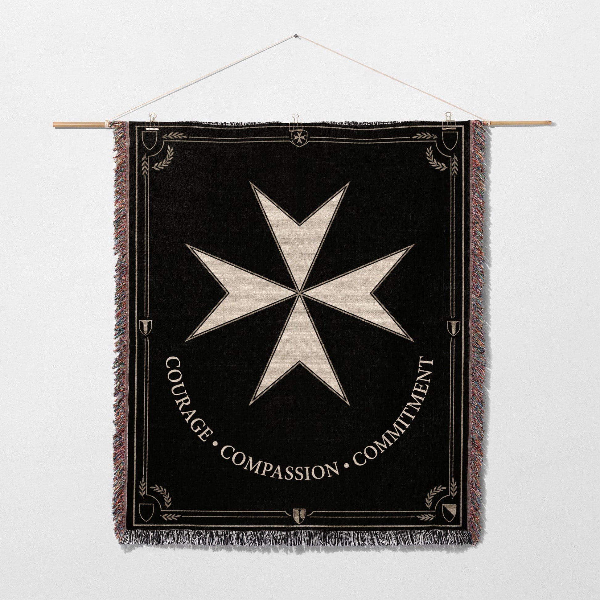 Black tapestry featuring a white Maltese cross and the words "COURAGE • COMPASSION • COMMITMENT".
