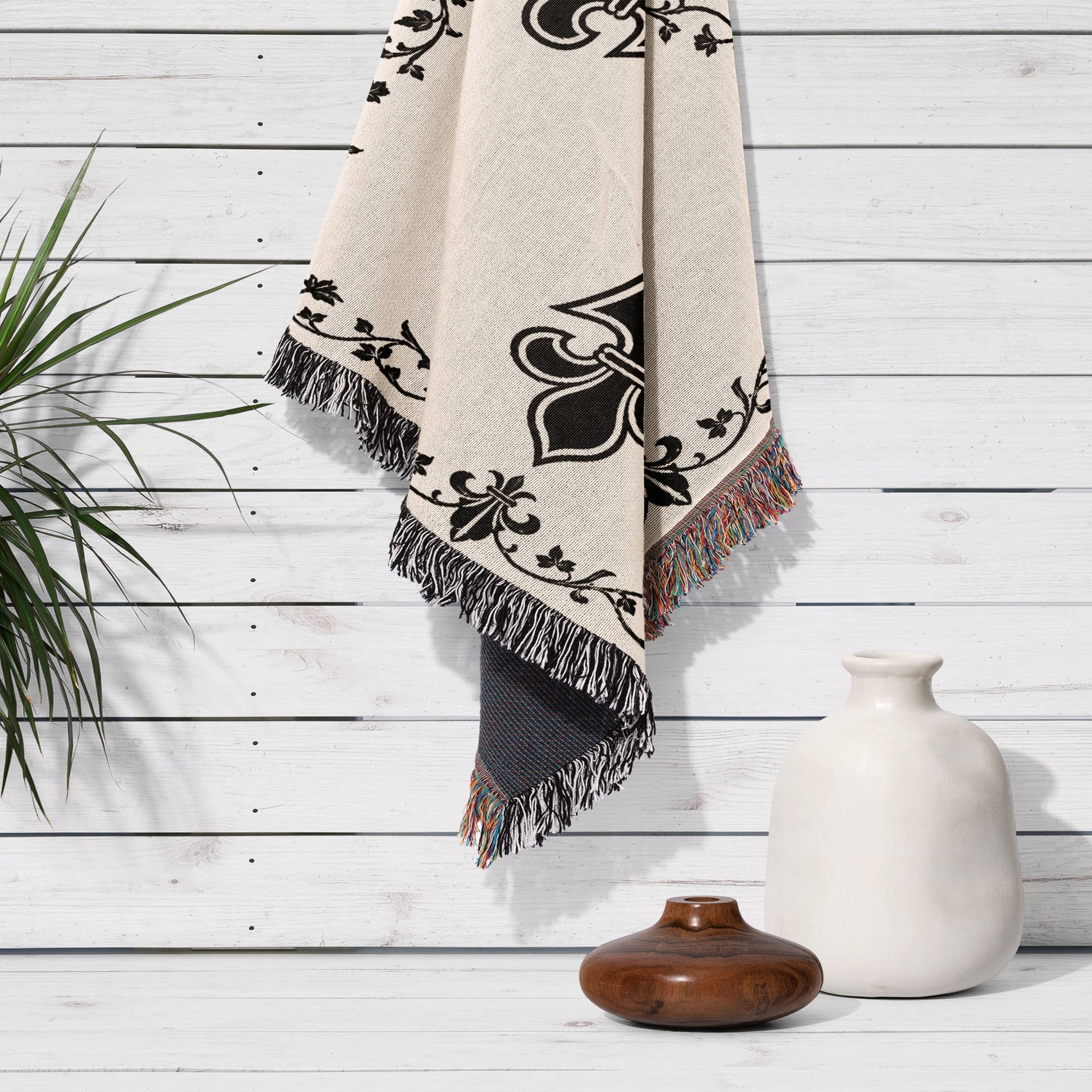 Cream blanket with black fleur-de-lis pattern and tasseled edges on wall.