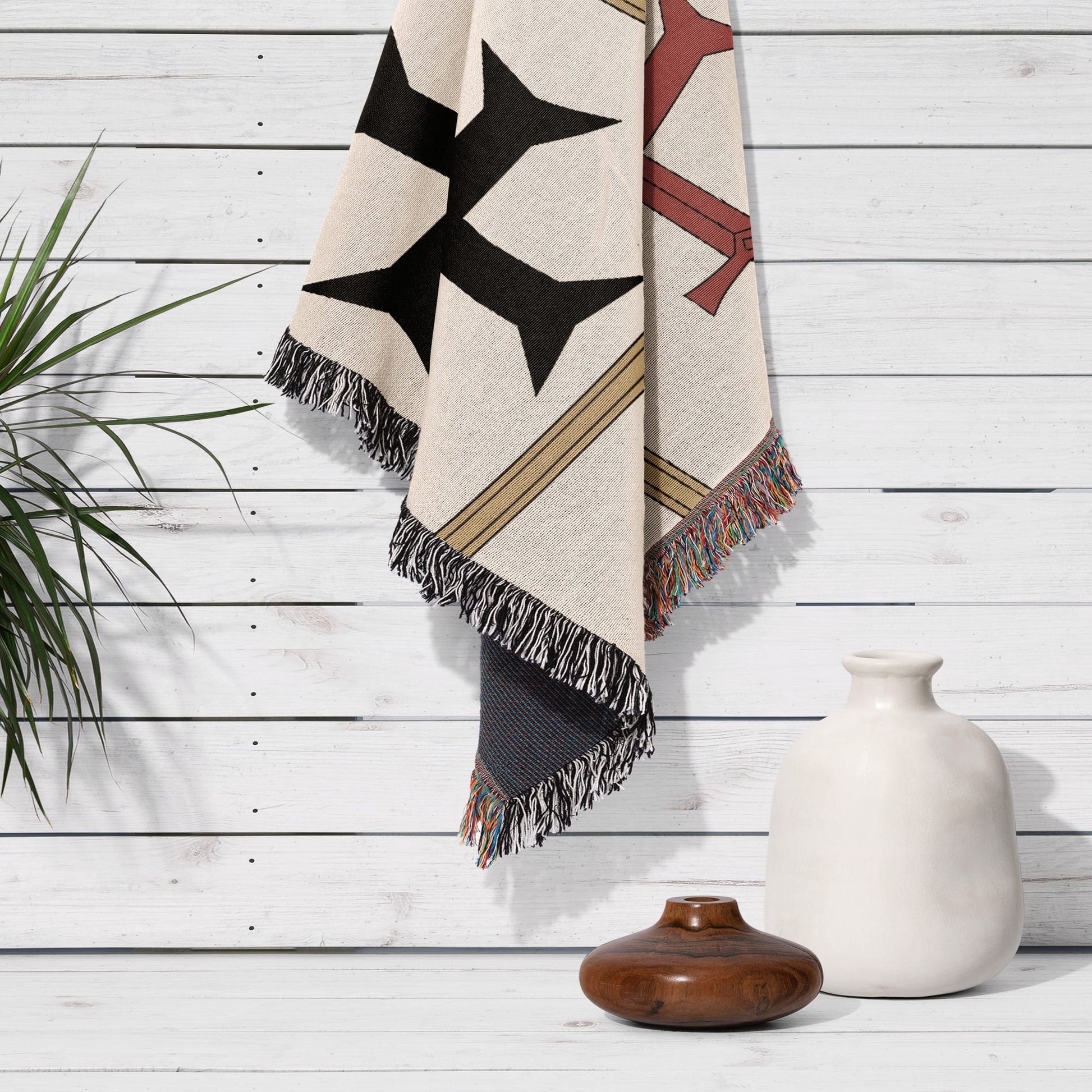 Textile blanket with geometric pattern and fringed edges draped over a white wooden wall.