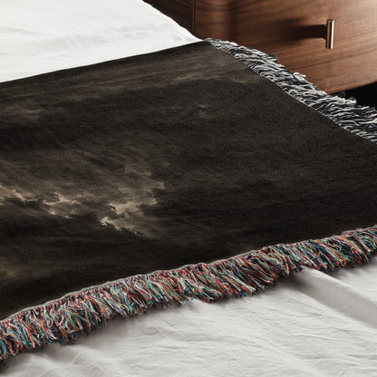 A dark woven throw blanket with a cloudy sky graphic and colorful fringe lies on a white bed.