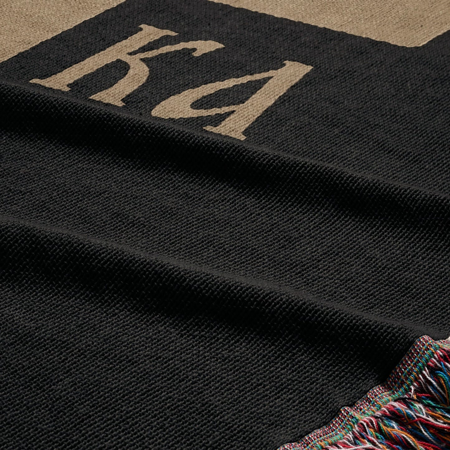 Black throw blanket with tan "KA" logo and striped fringe.