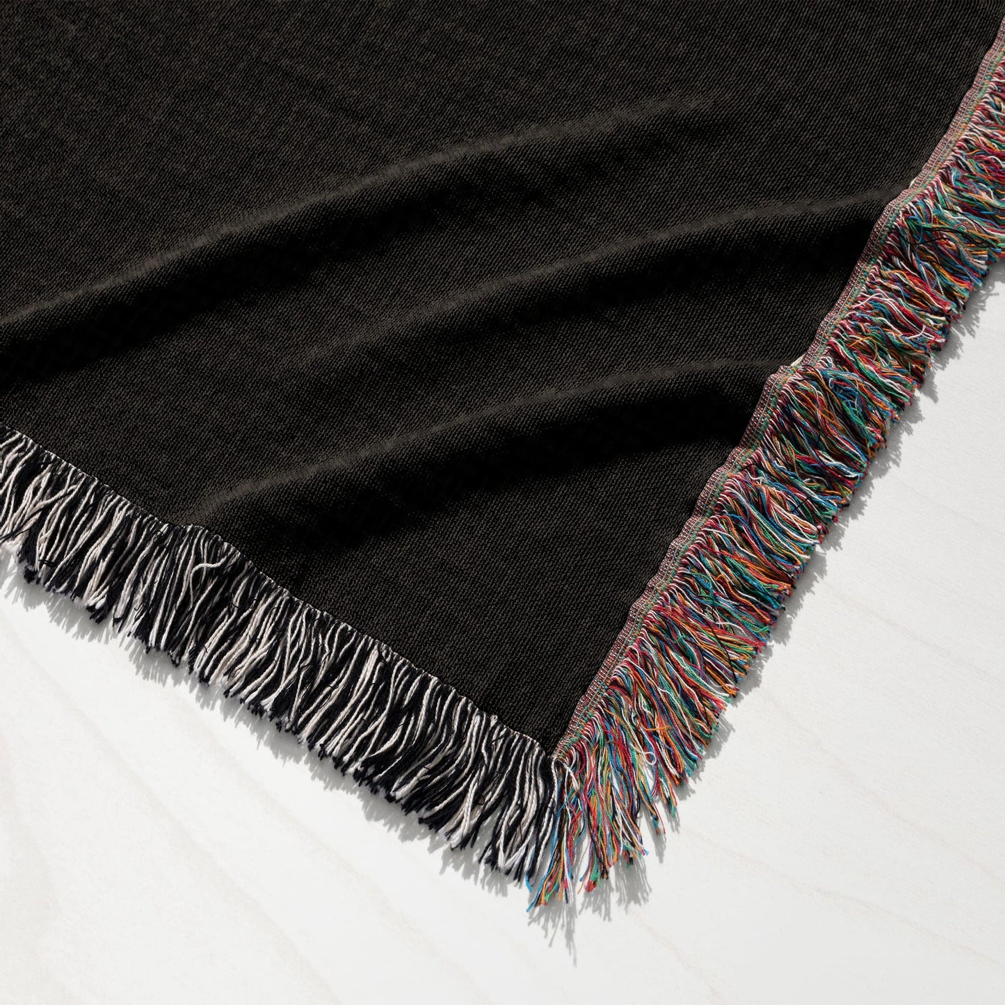 Black knit blanket with colorful fringed edges on a white wooden surface.