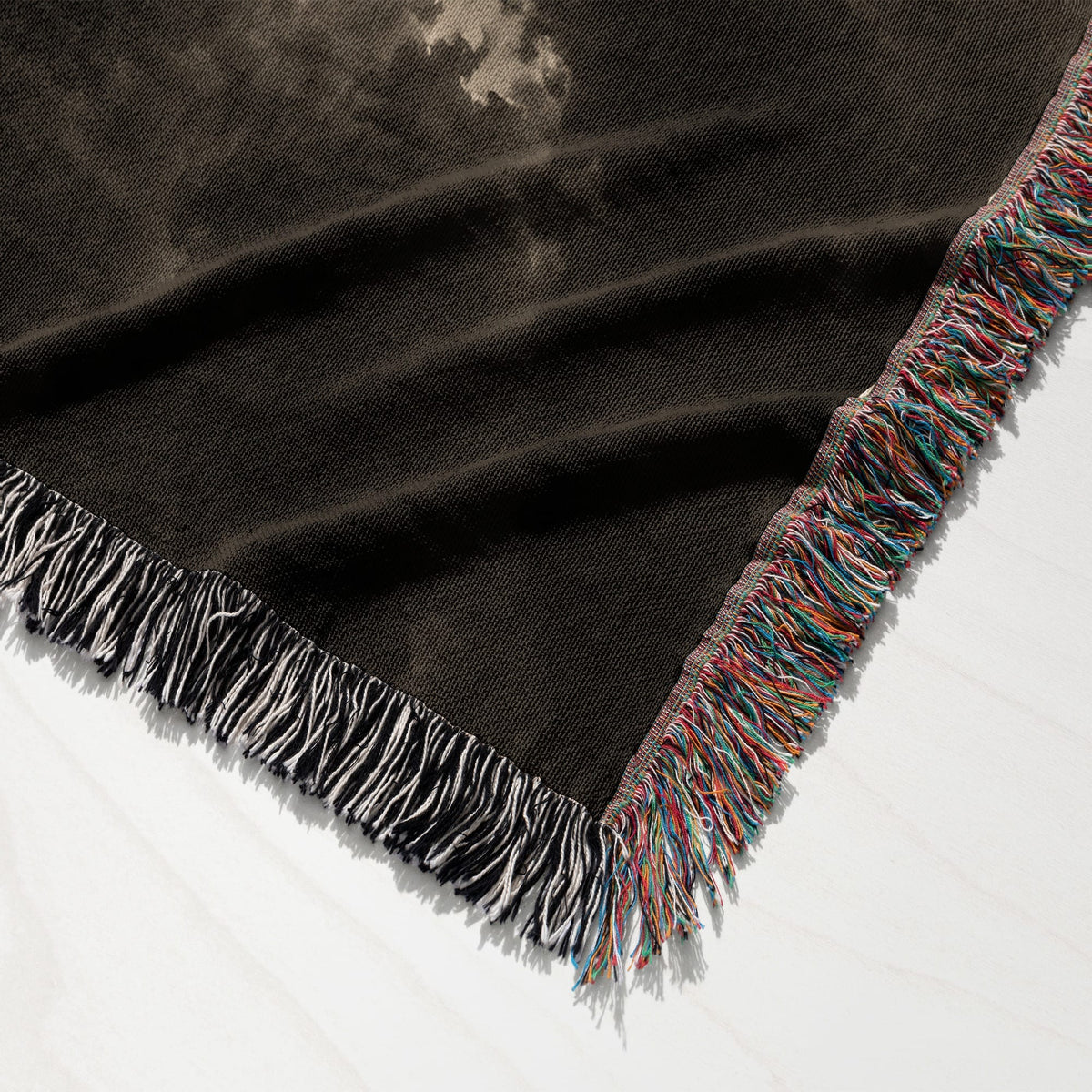 A dark woven throw blanket with a muted pattern, featuring colorful multicolored fringe along the edge.