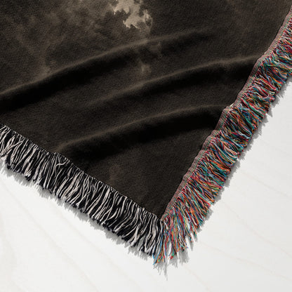 A dark woven throw blanket with a muted pattern, featuring colorful multicolored fringe along the edge.