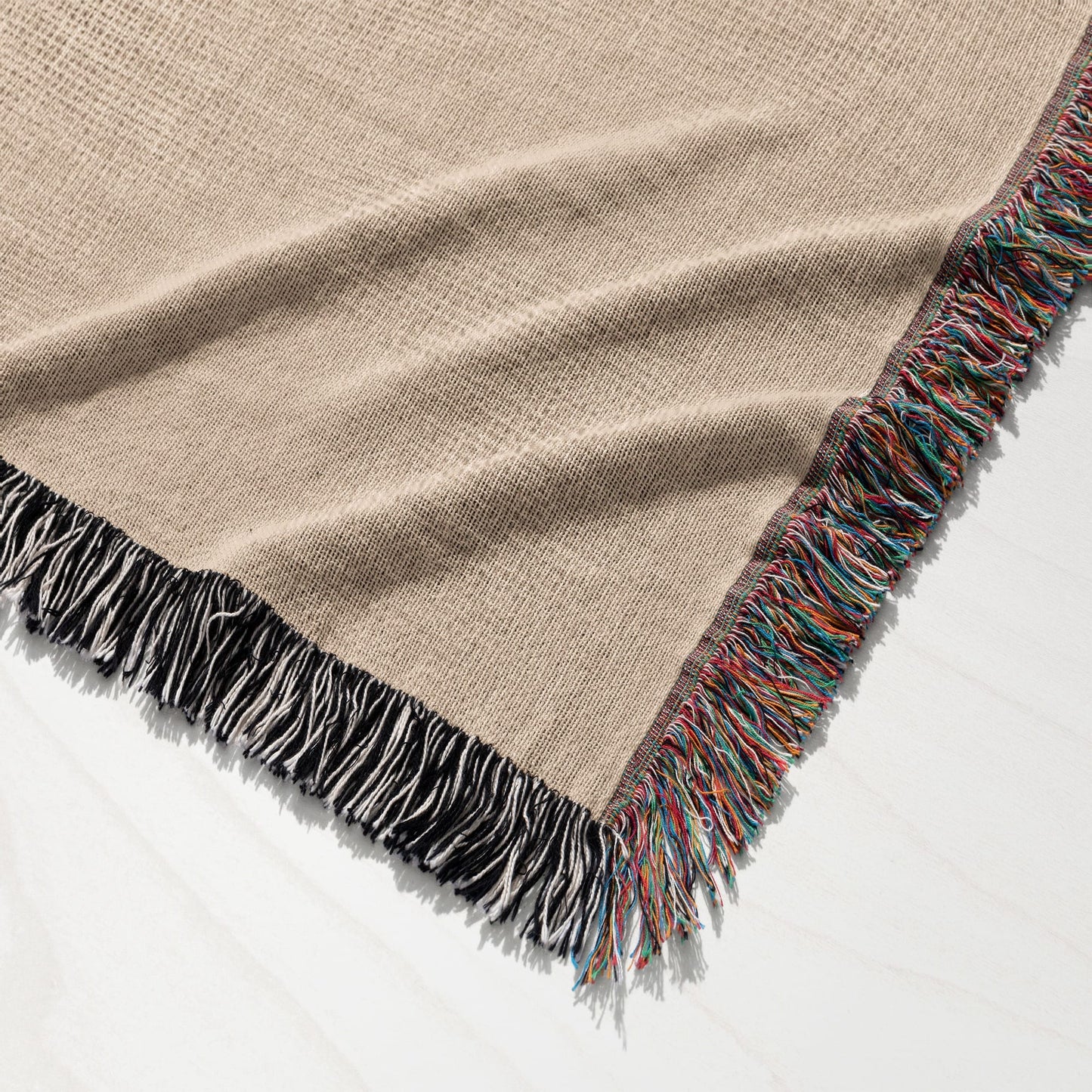 Tan woven blanket with colorful fringe detail on a white wooden surface.