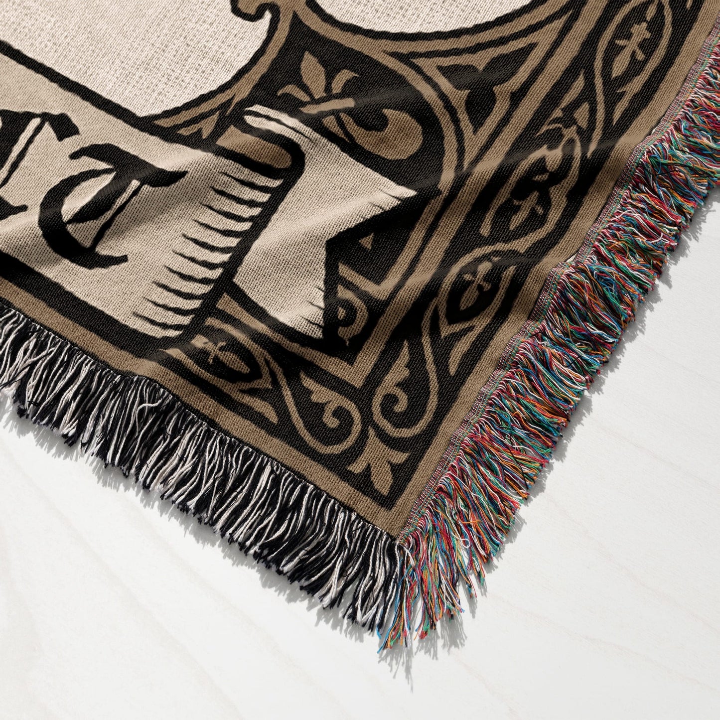Woven blanket with tasseled edges displaying black and tan ornate scrolling pattern and letters.
