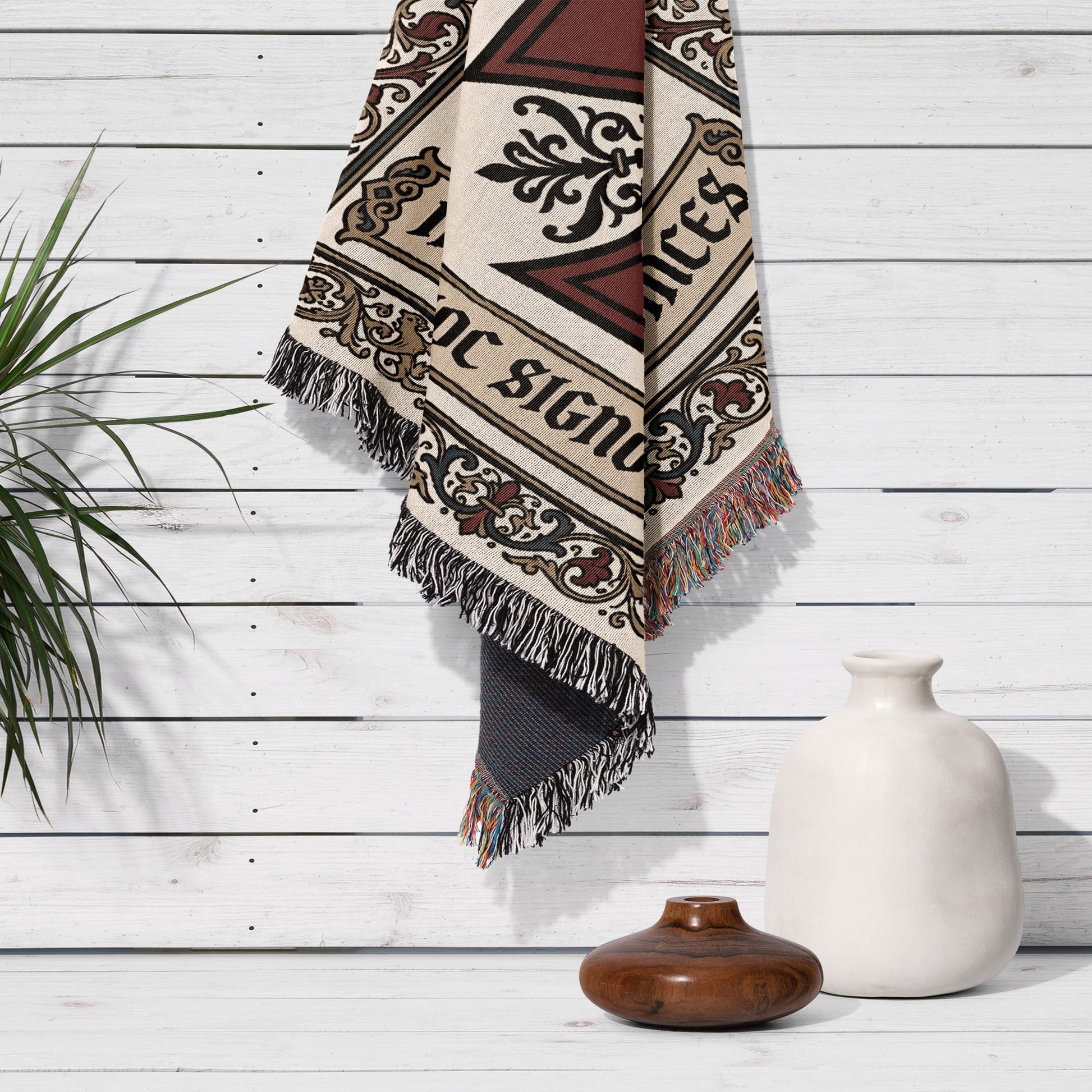 Woven blanket with fringe hanging on white wood wall next to vase.